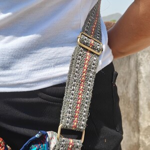 Pode incluir: Uma al&ccedil;a de bolsa transversal feita de tecido cinza com finas listras verticais vermelhas e amarelas, apresentando um ajustador de metal em tom de lat&atilde;o e um conector em D preso a uma bolsa de tecido acolchoado multicolorido com costuras verdes.