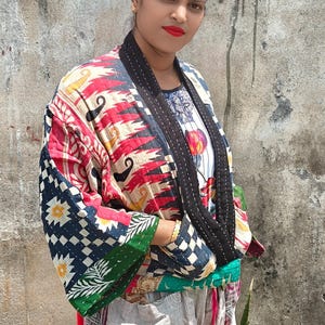 Può includere: Una giacca trapuntata caratterizzata da un vivace patchwork di motivi rossi, neri, bianchi e verdi con un colletto nero. Il capo presenta maniche larghe e un design multicolore con motivi floreali e geometrici, indossato sopra pantaloni chiari.