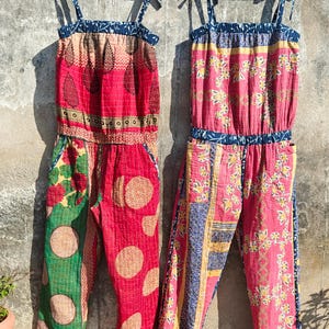 May include: Two colorful jumpsuits hanging on wooden hangers. The jumpsuits feature a patchwork design with red, pink, green, and blue fabrics. One has a leaf pattern, the other has floral and geometric patterns. Both have tie straps.