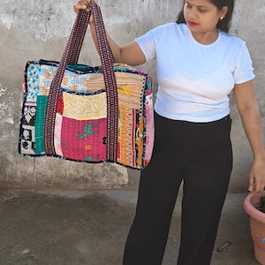 Puede incluir: Un bolso tote rectangular grande confeccionado con varios parches de tela coloridos que presentan diversos patrones, incluidos motivos animales. El bolso tiene dos asas largas de tela oscura estampada y costuras visibles en toda la superficie acolchada.