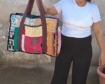 Pode incluir: Uma bolsa tote retangular grande feita de vários retalhos de tecido colorido com padrões diversos, incluindo motivos de animais. A bolsa possui duas alças longas de tecido escuro estampado e costuras visíveis em toda a superfície acolchoada.