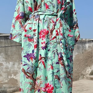 Può includere: Un accappatoio verde menta con una vivace stampa floreale e di uccelli. L'accappatoio presenta un design avvolgente con una cintura abbinata, tasche e maniche lunghe. La stampa include elementi rossi, viola e marroni.