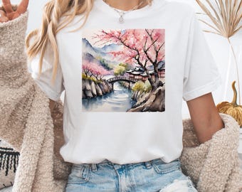 Pode incluir: Camiseta branca com uma ilustração em aquarela de uma paisagem japonesa com uma ponte, um rio e árvores de cerejeira em flor.