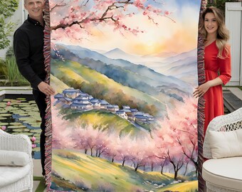 May include: A woven tapestry throw blanket features a watercolour painting of a village nestled in a valley with pink cherry blossoms and rolling green hills. The tapestry has a fringe border.