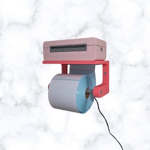 Puede incluir: Una impresora de etiquetas rosa está montada en un estante sobre un rollo de papel blanco y azul. Un cable negro se extiende desde la impresora. El fondo presenta un patrón de mármol.