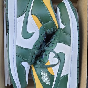 Puede incluir: Par de zapatillas altas verdes, blancas y amarillas en una caja de cart&oacute;n. Las zapatillas presentan una entresuela blanca, revestimientos verdes y un panel de tal&oacute;n amarillo. El logotipo de Nike es visible en el lateral.