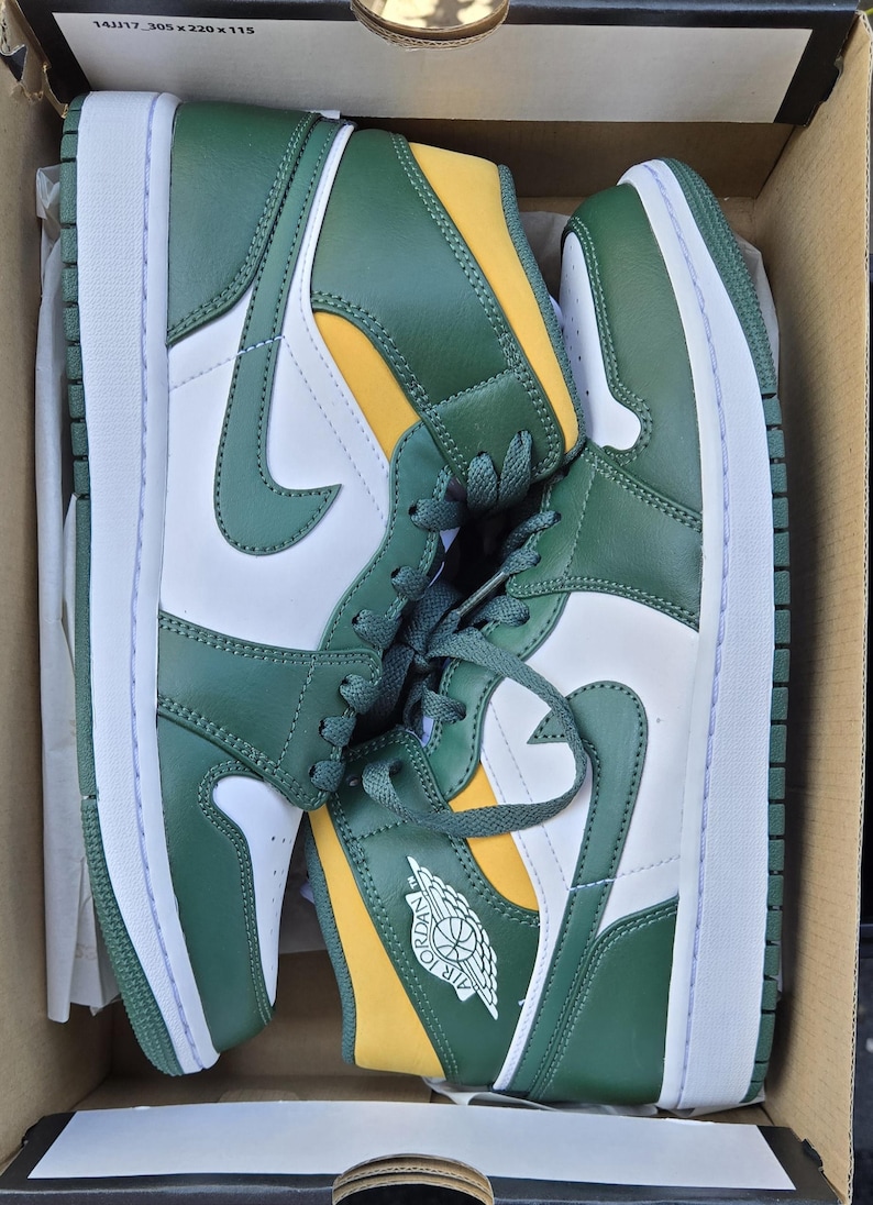 Puede incluir: Par de zapatillas altas verdes, blancas y amarillas en una caja de cart&oacute;n. Las zapatillas presentan una entresuela blanca, revestimientos verdes y un panel de tal&oacute;n amarillo. El logotipo de Nike es visible en el lateral.