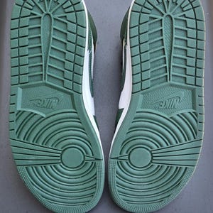 Puede incluir: Par de zapatillas verdes y blancas, vistas desde las suelas. Las suelas son de goma verde texturizada con el logo de Nike. Los zapatos tienen una entresuela blanca y cordones verdes.