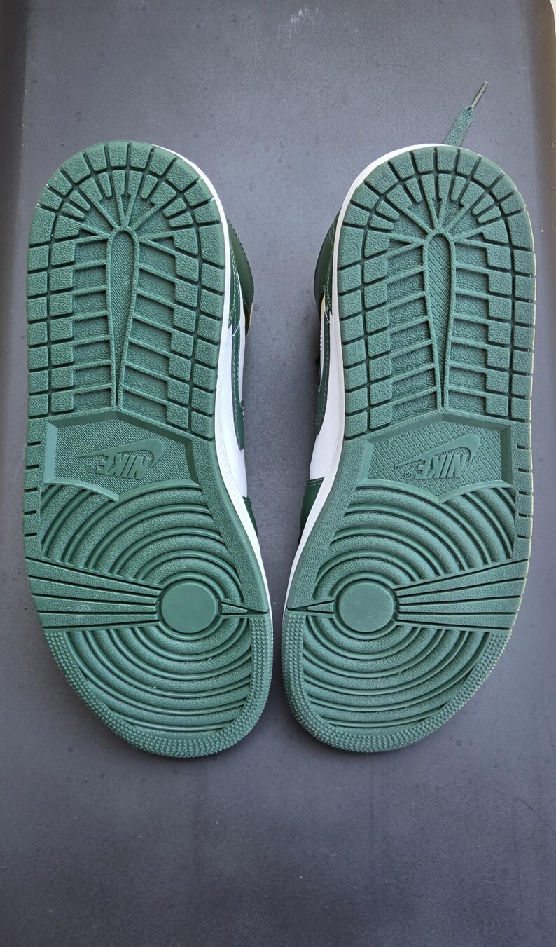 Puede incluir: Par de zapatillas verdes y blancas, vistas desde las suelas. Las suelas son de goma verde texturizada con el logo de Nike. Los zapatos tienen una entresuela blanca y cordones verdes.