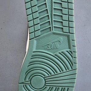 Puede incluir: Primer plano de la suela de una zapatilla deportiva verde y blanca. La suela est&aacute; texturizada con una variedad de patrones, incluido un logotipo de Nike, ranuras y un dise&ntilde;o circular. El zapato est&aacute; dise&ntilde;ado para la tracci&oacute;n y el agarre.