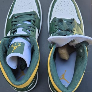 Puede incluir: Par de zapatillas bajas verdes, blancas y amarillas. Las zapatillas tienen la parte superior blanca con detalles en verde, cordones verdes y ribetes amarillos. La leng&uuml;eta de un zapato est&aacute; doblada, revelando el logotipo de Air Jordan.