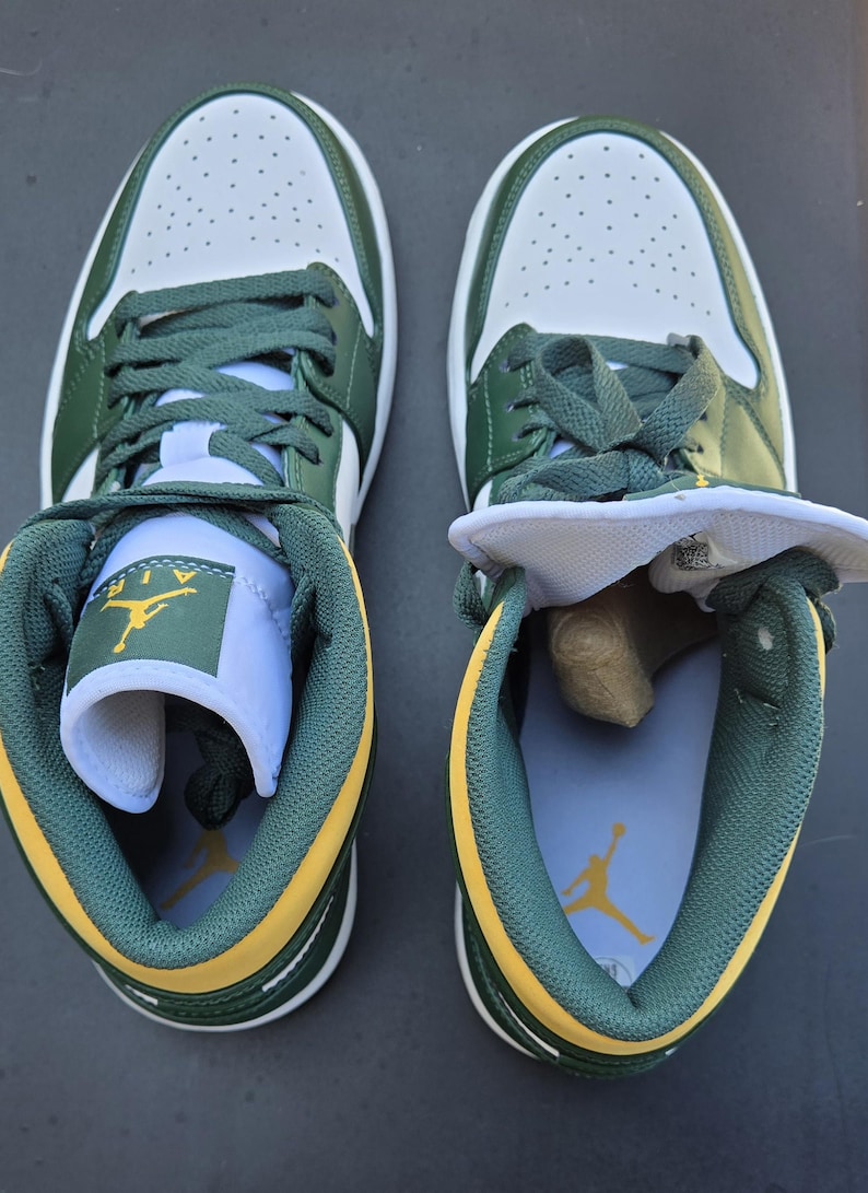 Puede incluir: Par de zapatillas bajas verdes, blancas y amarillas. Las zapatillas tienen la parte superior blanca con detalles en verde, cordones verdes y ribetes amarillos. La leng&uuml;eta de un zapato est&aacute; doblada, revelando el logotipo de Air Jordan.