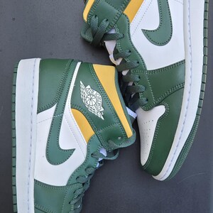 Puede incluir: Par de zapatillas altas verdes, blancas y amarillas. Las zapatillas presentan el logotipo de Air Jordan en el lateral y la leng&uuml;eta. Los zapatos tienen suelas blancas y cordones verdes. El fondo es negro liso.