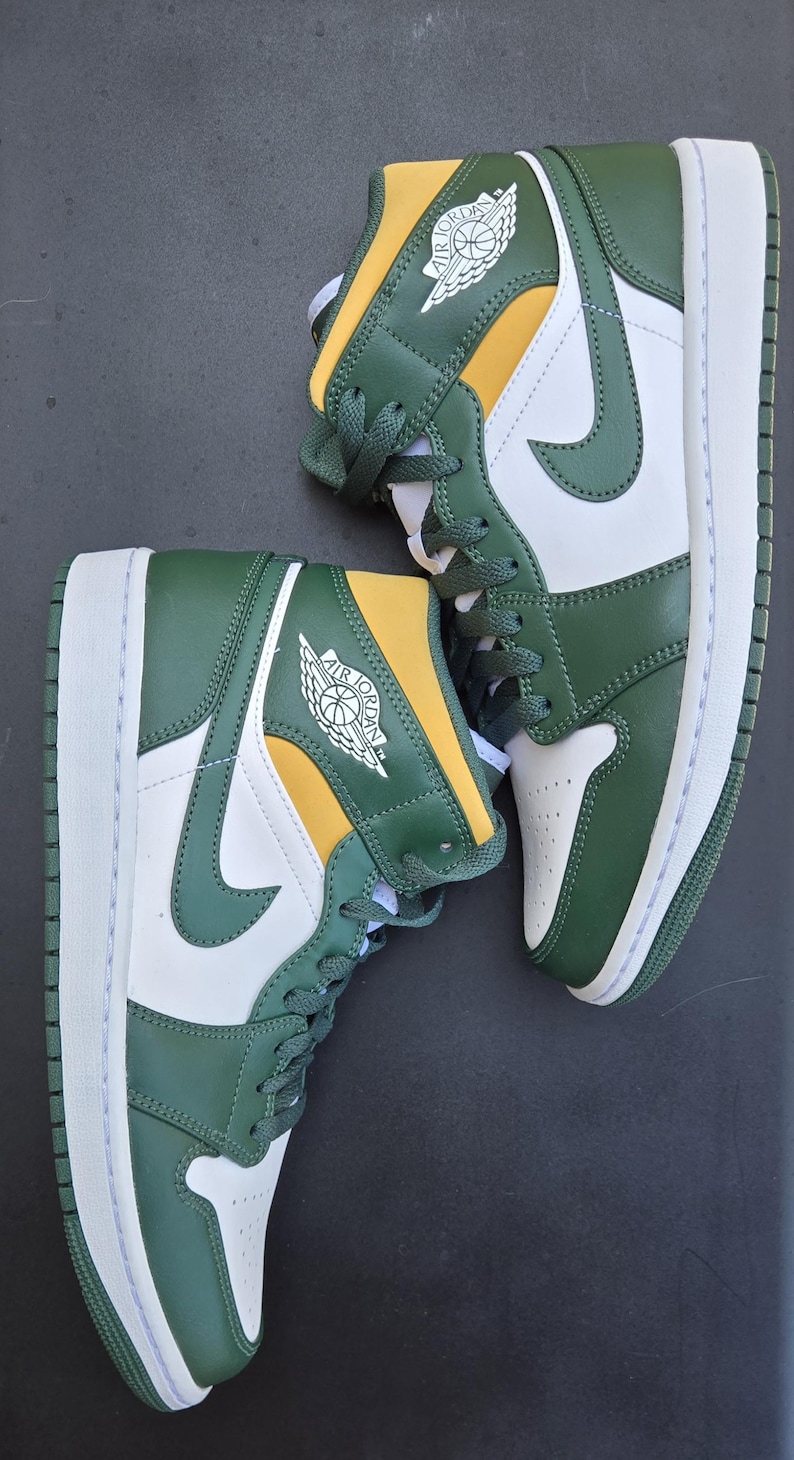 Puede incluir: Par de zapatillas altas verdes, blancas y amarillas. Las zapatillas presentan el logotipo de Air Jordan en el lateral y la leng&uuml;eta. Los zapatos tienen suelas blancas y cordones verdes. El fondo es negro liso.