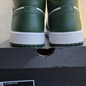 Puede incluir: Par de zapatillas Air Jordan 1 Mid verdes, blancas y amarillas. Las zapatillas est&aacute;n sobre una caja de zapatos negra. La caja tiene el texto "AIR JORDAN 1 MID" e informaci&oacute;n de tallas, incluyendo 27 cm.