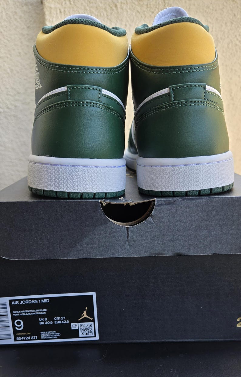 Puede incluir: Par de zapatillas Air Jordan 1 Mid verdes, blancas y amarillas. Las zapatillas est&aacute;n sobre una caja de zapatos negra. La caja tiene el texto "AIR JORDAN 1 MID" e informaci&oacute;n de tallas, incluyendo 27 cm.