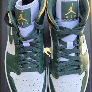 Puede incluir: Par de zapatillas altas verdes, blancas y amarillas. Las zapatillas presentan cordones verdes, un dise&ntilde;o verde y blanco y el logotipo de Air Jordan en amarillo. Las zapatillas est&aacute;n sobre una superficie negra.