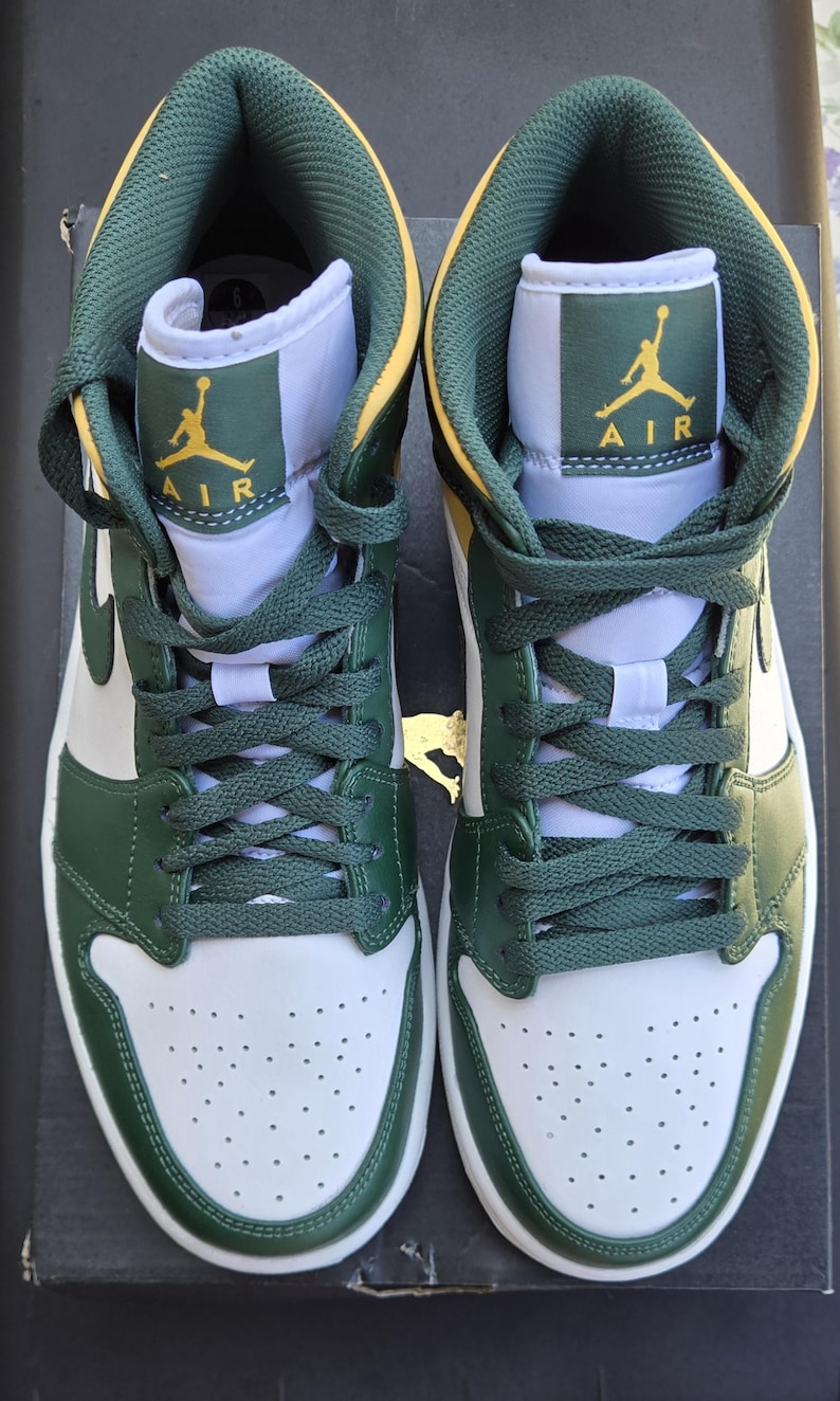 Puede incluir: Par de zapatillas altas verdes, blancas y amarillas. Las zapatillas presentan cordones verdes, un dise&ntilde;o verde y blanco y el logotipo de Air Jordan en amarillo. Las zapatillas est&aacute;n sobre una superficie negra.