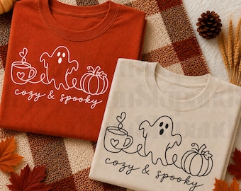 Puede incluir: Una camiseta de color óxido y una camiseta de color crema, cada una con un gráfico de líneas blancas o negras de una taza de café, un fantasma y una calabaza, con el texto "cozy & spooky" escrito en cursiva debajo de las ilustraciones.
