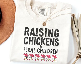 May include: Cream-colored t-shirt with the text "RAISING CHICKENS & FERAL CHILDREN" in black, distressed font. Below the text are cartoon chicken heads with red combs and googly eyes. A gold necklace is draped over the shirt.