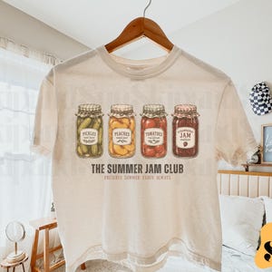 May include: Cream-colored t-shirt featuring a graphic of four jam jars labeled "Pickles", "Peaches", "Tomatoes", and "Strawberry Jam". The text "THE SUMMER JAM CLUB" and "PRESERVE SUMMER ENJOY ALWAYS" are printed below the jars.