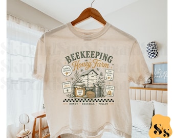 Puede incluir: Una camiseta beige de manga corta con un estampado gráfico que incluye el texto BEEKEEPING Honey Farm. El diseño muestra una colmena, un tarro de miel, un ahumador y un panal, rodeados de elementos florales y etiquetas con Raw & Unfiltered.