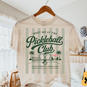 May include: Cream-colored t-shirt with a green and white graphic. The graphic reads "Meet Me At The Pickleball Club" with illustrations of palm trees, pickleball paddles, and a net. The shirt is hanging on a wooden hanger.