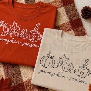 May include: A rust-colored t-shirt with white line art of a pumpkin, leaves, and a coffee mug with the text pumpkin season, and a beige t-shirt with black line art of the same design, both featuring a continuous line drawing style.