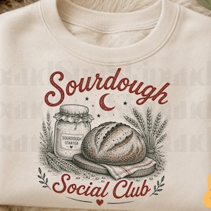 May include: A cream-coloured crewneck sweatshirt featuring a graphic illustration of a sourdough loaf, a jar labelled Sourdough Starter, and wheat stalks. The text Sourdough Social Club is printed in red, with a crescent moon and stars above the bread.