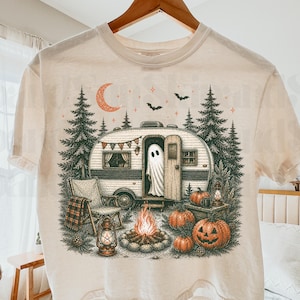 May include: A cream-colored t-shirt featuring a graphic illustration of a vintage camper in a forest setting. A ghost stands in the open doorway, with a campfire, pumpkins, lanterns, pine trees, a crescent moon, and bats surrounding the camper.