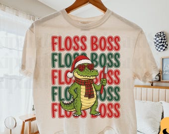 Puede incluir: Una camiseta color crema con el gráfico de un caimán verde que lleva un gorro de Papá Noel rojo y blanco, gafas de sol en forma de corazón y una bufanda de cuadros roja y verde. El caimán sostiene un cepillo de dientes. El texto FLOSS BOSS.