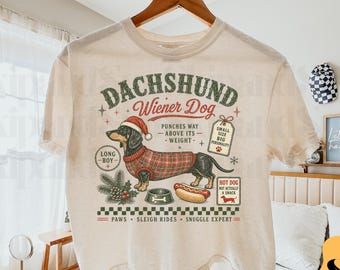 Puede incluir: Una camiseta de manga corta color crema con un gráfico de un perro salchicha con gorro de Papá Noel y un suéter de cuadros rojos. El diseño incluye textos como DACHSHUND, Wiener Dog, LONG BOY, y Paws, Sleigh Rides, Snuggle Expert, con un icono de perrito caliente.