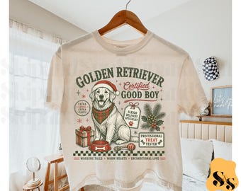 Puede incluir: Una camiseta beige con un gráfico de un Golden Retriever con gorro de Papá Noel y pañuelo a cuadros. El diseño incluye una caja de regalo, un cuenco, una rama de pino y el texto Golden Retriever, Certified Good Boy, Sleigh Delivery Specialist y Professional Treat Tester.