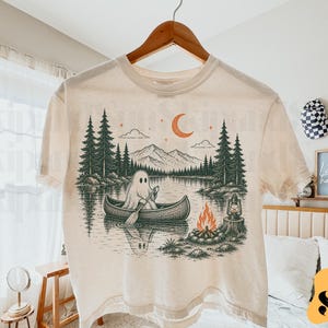 May include: A cream-coloured short-sleeve t-shirt featuring a graphic illustration of a ghost paddling a canoe on a lake. The scene includes dark green pine trees, mountains, a crescent moon, a campfire, and a lantern on a tree stump.