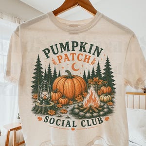 May include: A cream-colored short-sleeve t-shirt featuring a graphic print of pumpkins, a campfire, a lantern, and pine trees. The text PUMPKIN PATCH SOCIAL CLUB is printed in dark green and orange, with a crescent moon icon above the pumpkins.
