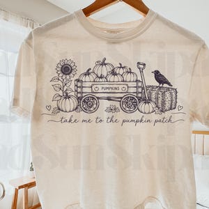 Może przedstawiać: Kremowy t-shirt z krótkim rękawem z czarną grafiką liniową przedstawiającą drewniany wóz wypełniony dyniami, słonecznikiem, wroną i belą siana. Napis "PUMPKINS" oraz "take me to the pumpkin patch" jest nadrukowany w kolorze czarnym.