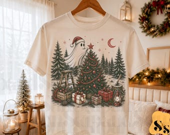 Puede incluir: Una camiseta de color crema con una ilustración gráfica de un fantasma con gorro de Papá Noel colocando una estrella en un árbol de Navidad. La escena incluye pinos, regalos envueltos, una escalera de madera, un farol y una luna creciente.