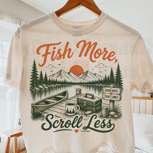 May include: A cream-colored t-shirt featuring a graphic print of a lake scene with a canoe, a tackle box, a coffee mug, and a sign that reads "GOOD DAYS TIGHT LINES". The text "Fish More," and "Scroll Less" is printed in orange and dark green.