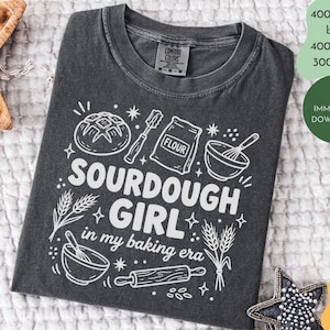 Może przedstawiać: Ciemnoszara koszulka z białym nadrukiem. Projekt zawiera ilustracje związane z pieczeniem i tekst "SOURDOUGH GIRL in my baking era." Zawiera bochenek chleba, worek mąki, trzepaczkę i wałek.