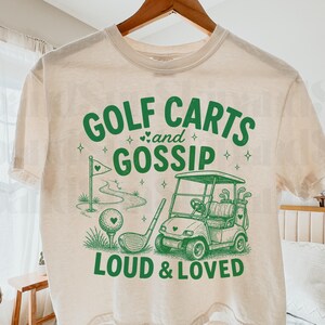 以下が含まれることがあります： 「GOLF CARTS and GOSSIP LOUD & LOVED」と書かれた緑色のグラフィックが特徴のクリーム色のTシャツ。グラフィックには、ゴルフカート、ゴルフクラブ、ゴルフボール、旗のあるゴルフコースが描かれています。木製のハンガーにTシャツが掛かっています。