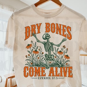 May include: A cream-colored short-sleeve t-shirt featuring a graphic of a skeleton kneeling among orange flowers. The text reads DRY BONES COME ALIVE EZEKIEL 37:5 in bold orange lettering. The skeleton has its arms raised upward toward the text.