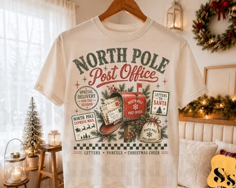Puede incluir: Una camiseta de color crema con un estampado gráfico de un buzón rojo con la etiqueta North Pole Post Office, rodeado de ramas de pino, cartas y un saco. El texto dice North Pole Post Office, Special Delivery y Letters to Santa.