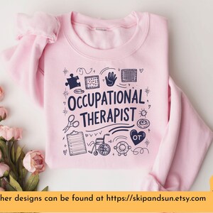Puede incluir: Sudadera rosa con el texto "Occupational Therapist" en letras azul marino. El dise&ntilde;o incluye varios iconos relacionados con la terapia ocupacional, como una pieza de rompecabezas, una mano, una silla de ruedas y un estetoscopio.