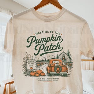 May include: A cream-colored t-shirt featuring a graphic print with the text "Meet me by the Pumpkin Patch" in dark green script. The illustration depicts an orange pickup truck loaded with pumpkins, a wooden sign, and a barn in a rural setting.