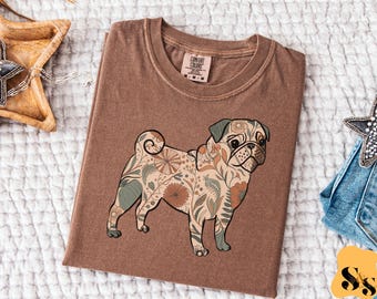 May include: Brown Comfort Colors t-shirt featuring a floral patterned pug design. The pug is tan, green, and brown. The shirt has a crew neck and short sleeves. The tag reads Comfort Colors.