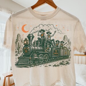 May include: A cream-coloured t-shirt featuring a graphic of a steam locomotive in a desert landscape. Four skeletons wearing cowboy hats ride the train. The text reads Good Times Est. 1910 and Ride Forever. An orange crescent moon and stars are above.