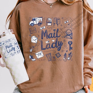Peut inclure: Un sweat-shirt marron avec un imprim&eacute; graphique bleu et blanc de bo&icirc;tes aux lettres, d'enveloppes et d'un camion de livraison. Le texte "Mail Lady" est imprim&eacute; en bleu sur le devant du sweat-shirt.