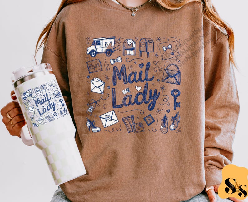 Peut inclure: Un sweat-shirt marron avec un imprim&eacute; graphique bleu et blanc de bo&icirc;tes aux lettres, d'enveloppes et d'un camion de livraison. Le texte "Mail Lady" est imprim&eacute; en bleu sur le devant du sweat-shirt.