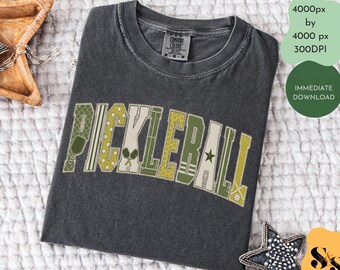 Puede incluir: Una camiseta de manga corta color gris carbón con cuello redondo. El frente presenta la palabra PICKLEBALL en letras grandes de estilo bloque, cada una rellena con patrones en tonos verdes, crema y amarillos, incluyendo estrellas y paletas.