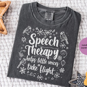 May include: Dark gray t-shirt with white text and illustrations. The shirt features the words "Speech Therapy helps little voices take flight" with bird and snowflake designs. A round purple sticker says "Immediate Download".